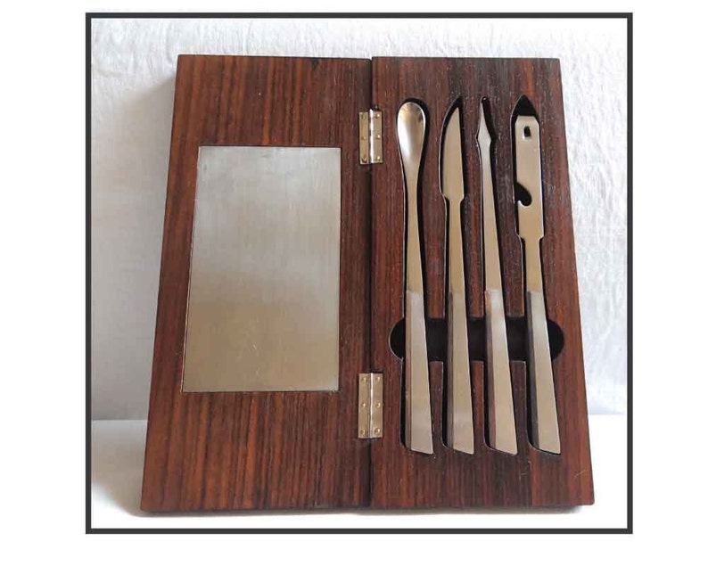 Peut inclure: Une bo&icirc;te en bois avec un couvercle &agrave; charni&egrave;res, contenant cinq outils en argent. La bo&icirc;te est en bois fonc&eacute; avec un motif de grain. Les outils sont dispos&eacute;s en rang&eacute;e, les poign&eacute;es vers le haut. Les outils sont de formes et de tailles diff&eacute;rentes, et ils semblent &ecirc;tre en m&eacute;tal.