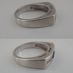 Op de afbeelding: Een zilveren ring met een blauwe edelsteen en een rij kleinere, heldere edelstenen. De ring heeft een vierkante band met een ontwerp van drie lijnen.