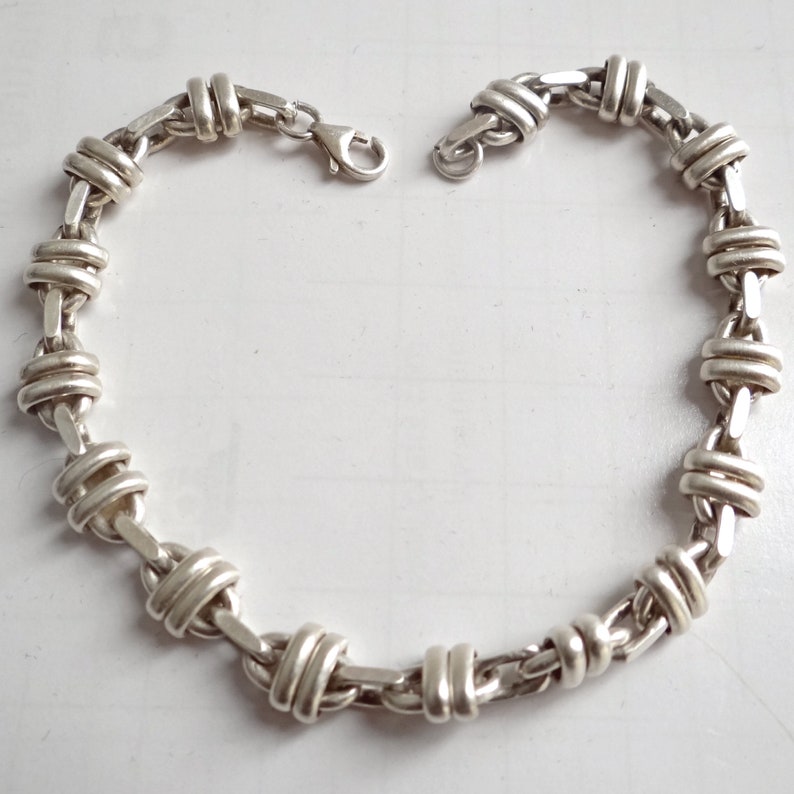 May include: A silver chain bracelet with a unique, interwoven design. The links are thick and have a polished finish.
