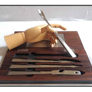 Peut inclure: Un mod&egrave;le de main en bois tient un couteau et une cuill&egrave;re en argent sur un plateau en bois fonc&eacute; avec quatre autres couteaux en argent dans des fentes.