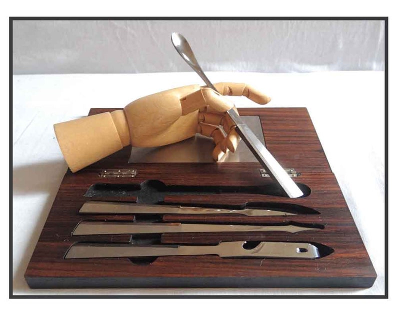 Peut inclure: Un mod&egrave;le de main en bois tient un couteau et une cuill&egrave;re en argent sur un plateau en bois fonc&eacute; avec quatre autres couteaux en argent dans des fentes.