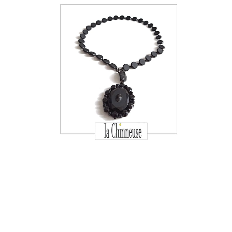 May include: A black necklace with a large black pendant. The pendant is a round, faceted, black glass button with a smaller black glass button in the center. The necklace is made of black glass beads.