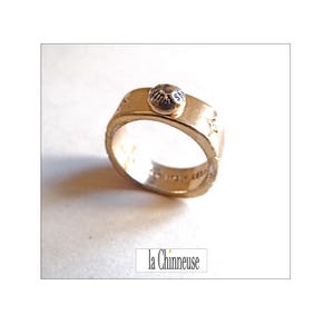 Könnte beinhalten: Ein goldfarbener Ring mit strukturiertem Band und einem kleinen, runden, silberfarbenen Schmuckelement. Der Ring hat sternförmige Details und der Text "la Chinneuse" ist im Bild sichtbar.