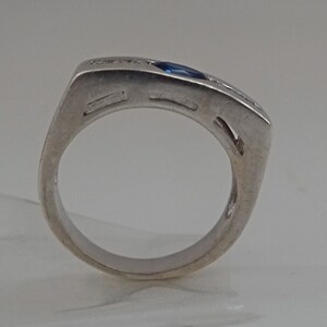Op de afbeelding: Een zilveren ring met een blauwe edelsteen en kleine heldere stenen gezet in een vierkante band.