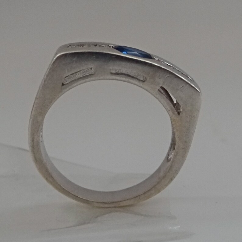 Op de afbeelding: Een zilveren ring met een blauwe edelsteen en kleine heldere stenen gezet in een vierkante band.