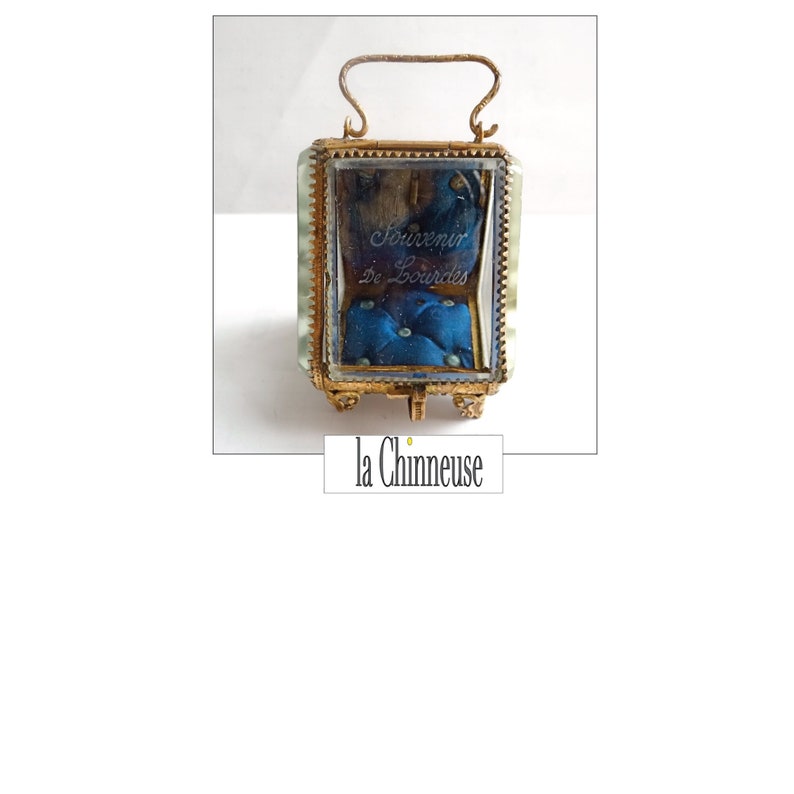 Op de afbeelding: Een kleine, vierkante glazen doos met een gouden frame en een blauwe fluwelen binnenkant. De doos heeft een handvat aan de bovenkant en is versierd met een gouden rand. De doos is gegraveerd met de woorden "Souvenir de Lourdes".