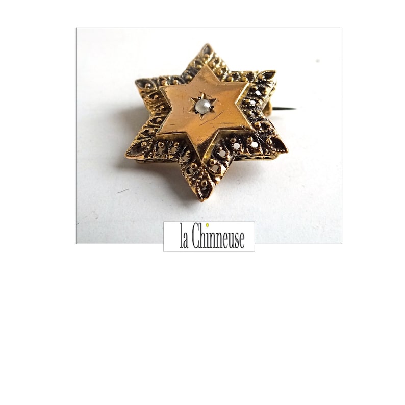 Op de afbeelding: Een gouden Davidster-broche met een parel in het midden. De broche is versierd met ingewikkelde filigraan details.