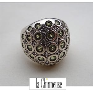 May include: A silver ring with a rounded top, covered in small, round, olive-green gemstones and tiny clear stones. The ring has a smooth, polished finish. The text "la Chinneuse" is at the bottom.