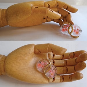 May include: Two gold-tone hoop earrings with pink and white floral designs. The earrings are held in a wooden hand.
