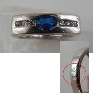 Op de afbeelding: Een zilveren ring met een blauwe ovale edelsteen en een rij kleinere heldere edelstenen. De ring is gemarkeerd met "925" aan de binnenkant.