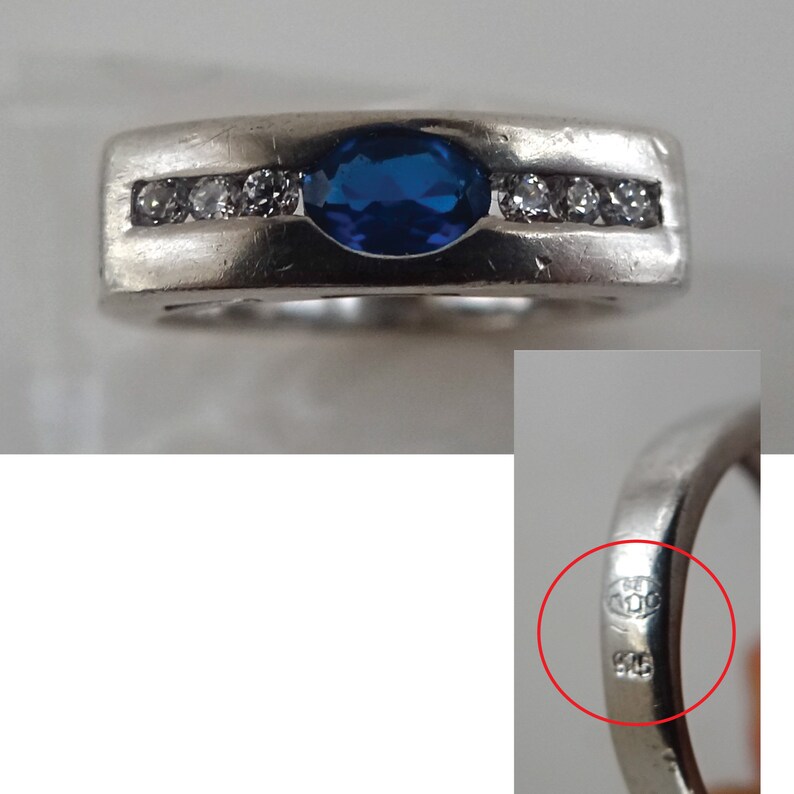 Op de afbeelding: Een zilveren ring met een blauwe ovale edelsteen en een rij kleinere heldere edelstenen. De ring is gemarkeerd met "925" aan de binnenkant.