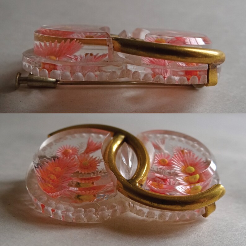 May include: A vintage brooch featuring two clear glass circles, each containing pink and yellow flowers. The circles are joined by a gold metal band.