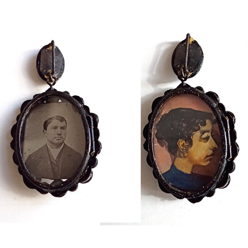 May include: Two oval-shaped locket pendants with black metal frames. One locket features a black and white photograph of a man with a mustache and beard. The other locket features a hand-painted portrait of a woman with dark hair and a blue dress.