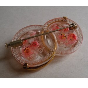 May include: A pink glass brooch with a floral design and a gold metal pin. The brooch is made of two interlocking circles.