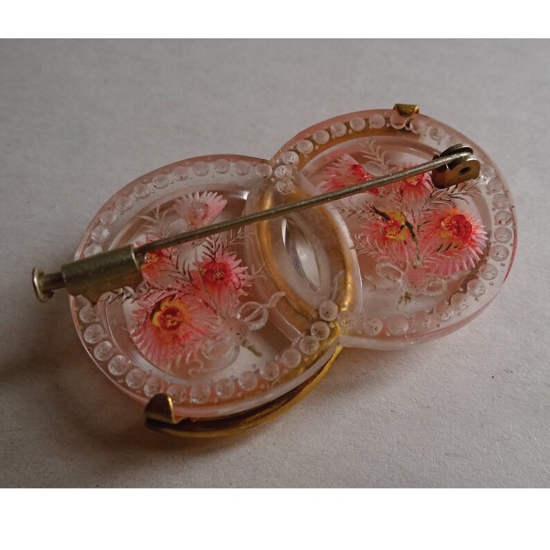 May include: A pink glass brooch with a floral design and a gold metal pin. The brooch is made of two interlocking circles.
