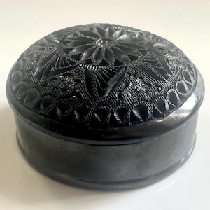 Può includere: Un portagioie nero e rotondo con un motivo floreale intagliato sul coperchio. La scatola è realizzata in un materiale scuro e lucido e ha una base liscia e arrotondata. Il coperchio presenta motivi e trame intricate.
