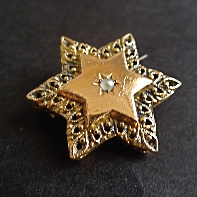 Op de afbeelding: Een gouden Davidster-broche met een parel in het midden. De ster is omgeven door een filigraanontwerp.
