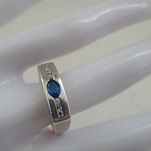 Op de afbeelding: Een zilveren ring met een blauwe ovale edelsteen en een rij heldere ronde edelstenen aan elke kant.