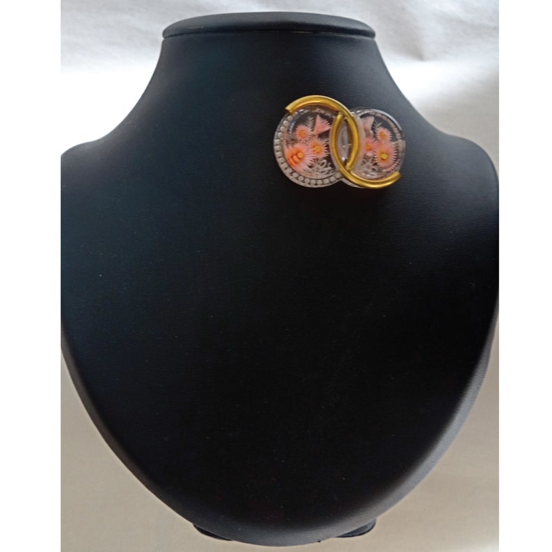 May include: A pair of gold and silver earrings with a floral design inside the clear plastic. The earrings are on a black display stand.