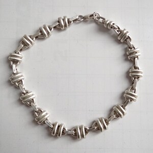 May include: A silver chain bracelet with a unique, interlocking design. The bracelet is made of a series of linked, oval-shaped pieces that are interwoven to create a secure and stylish look.