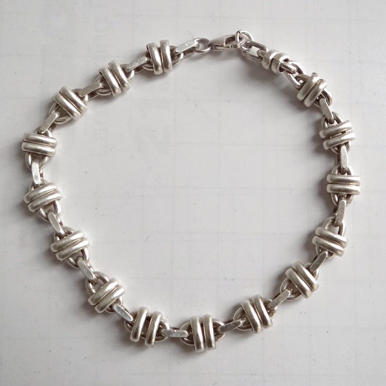 May include: A silver chain bracelet with a unique, interlocking design. The bracelet is made of a series of linked, oval-shaped pieces that are interwoven to create a secure and stylish look.