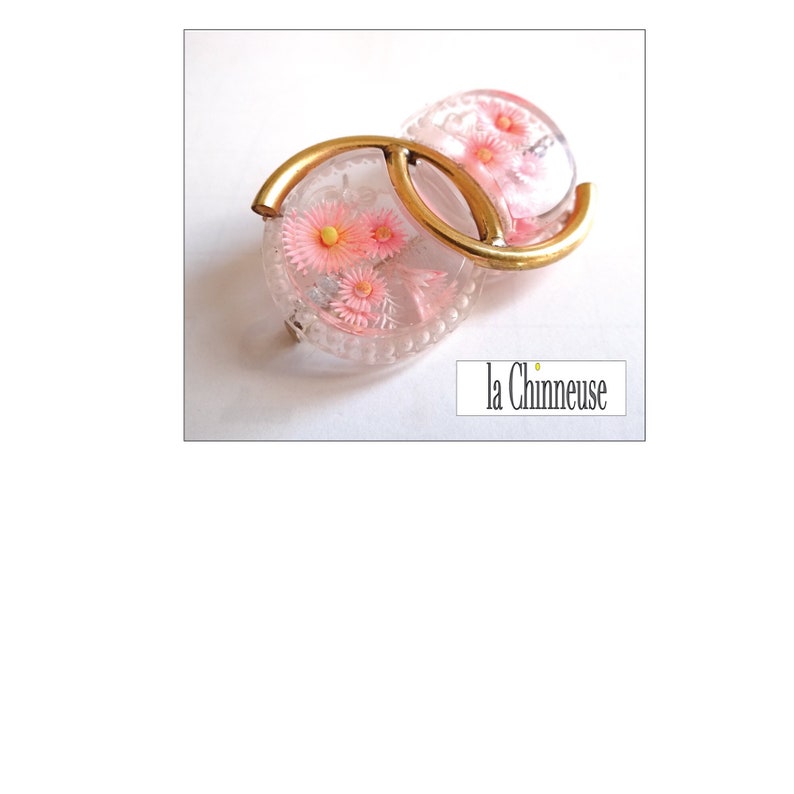 May include: A vintage brooch featuring a gold metal frame and two clear glass circles containing pink flowers. The brooch is shaped like the letter "C".