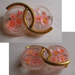May include: A clear plastic brooch with a gold metal frame. The brooch is shaped like two interlocking circles, each with a floral design. The flowers are pink with yellow centres.