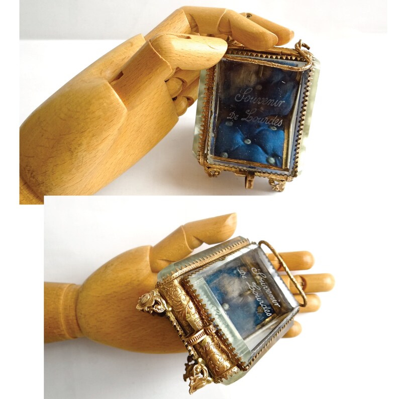 Op de afbeelding: Een klein, versierd, goudkleurig glazen doosje met een blauwe fluwelen binnenkant. Het doosje heeft een scharnierend deksel en is versierd met bloemmotieven. De inscriptie "Souvenir de Lourdes" is in het glas gegraveerd.