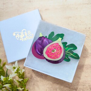May include: A purple and pink fig brooch with a green leaf design. The brooch is made of resin and is a whimsical and colorful accessory.