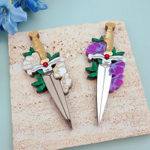 Könnte beinhalten: Zwei Broschen aus Acryl in Form von Dolchen mit floralen Akzenten. Ein Dolch hat weiße Blüten und grüne Blätter, der andere hat violette Blüten und grüne Blätter.