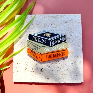 Pode incluir: Um conjunto de três broches de esmalte que representam a literatura clássica. O broche superior é preto com texto branco que diz "The Illiad". O broche do meio é creme com texto preto que diz "The Odyssey". O broche inferior é laranja com texto preto que diz "The Aeneid".