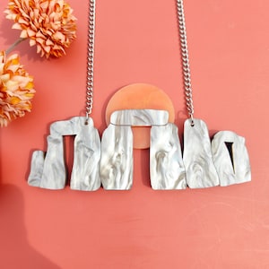 May include: A silver chain necklace with a white and grey stone pendant shaped like Stonehenge. The pendant is made of a marbled material and has a textured surface.