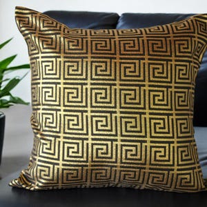 May include: A gold and black patterned throw pillow with a geometric design. The pillow is on a black leather couch.