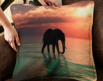 May include: A throw pillow with a silhouette of an elephant walking in the water at sunset. The background is a vibrant orange and pink sunset with clouds.