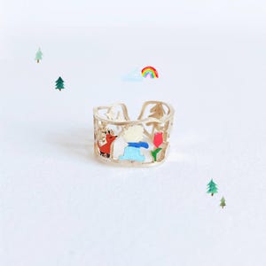 May include: A gold-toned open band ring featuring intricate cut-out details of a small red fox, a figure in a blue coat, and a red tulip. The design includes small star and tree shapes integrated into the metal band, creating a narrative scene.