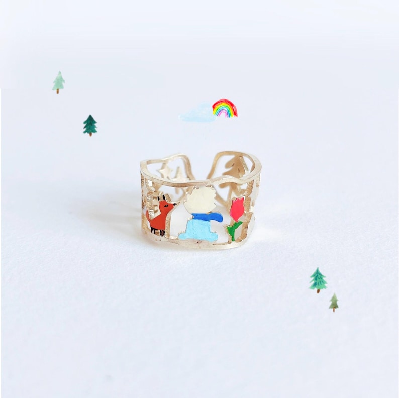 May include: A gold-toned open band ring featuring intricate cut-out details of a small red fox, a figure in a blue coat, and a red tulip. The design includes small star and tree shapes integrated into the metal band, creating a narrative scene.