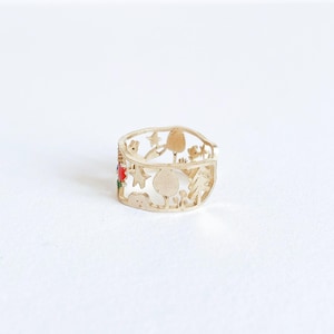 May include: A gold-toned metal ring featuring a cut-out design of various shapes including trees, stars, and rounded forms. A small section of the ring is accented with vibrant red and green enamel details, creating a contrast against the metallic finish.