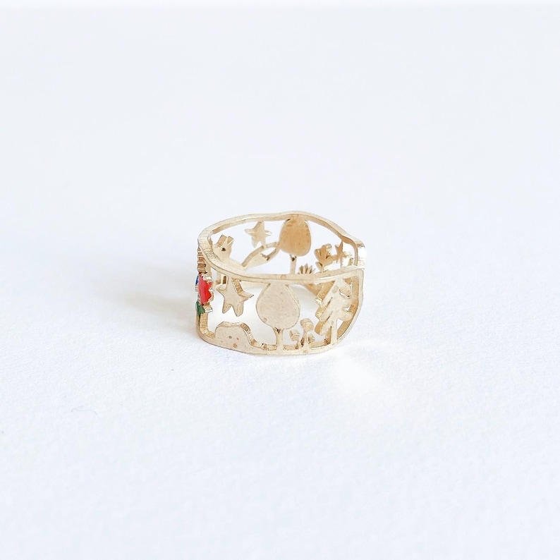 May include: A gold-toned metal ring featuring a cut-out design of various shapes including trees, stars, and rounded forms. A small section of the ring is accented with vibrant red and green enamel details, creating a contrast against the metallic finish.