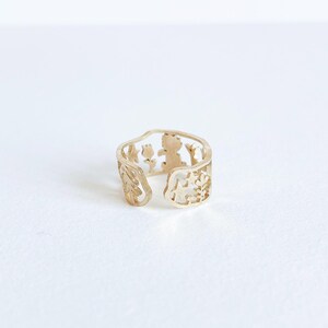 May include: A gold-toned open ring featuring a cut-out design that includes silhouettes of a small tree, a flower, a star, and a figure. The band has an irregular, organic shape with an opening at the front, showcasing a detailed metalwork pattern.