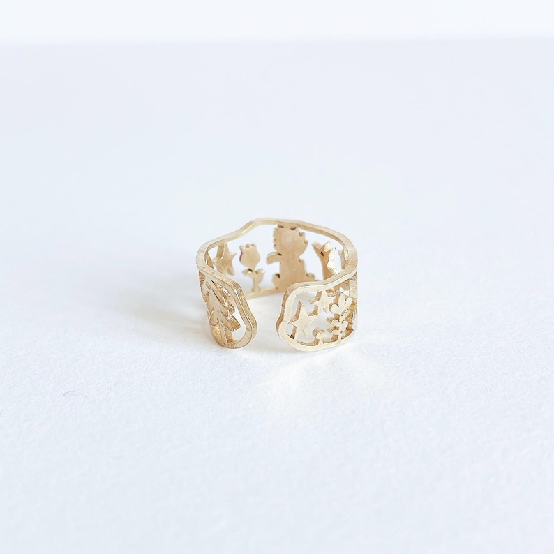 May include: A gold-toned open ring featuring a cut-out design that includes silhouettes of a small tree, a flower, a star, and a figure. The band has an irregular, organic shape with an opening at the front, showcasing a detailed metalwork pattern.