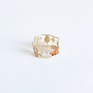 May include: A gold-toned metal ring featuring a cut-out design of a forest scene. The band displays silhouettes of trees, stars, and a small fox with orange enamel detailing. The ring has an openwork structure with a smooth, polished finish.