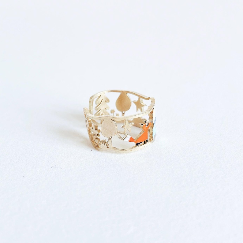 May include: A gold-toned metal ring featuring a cut-out design of a forest scene. The band displays silhouettes of trees, stars, and a small fox with orange enamel detailing. The ring has an openwork structure with a smooth, polished finish.