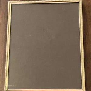 May include: A rectangular gold-colored picture frame with a dark brown interior. The frame has a detailed rope-like pattern around the edges. The frame is sitting on a dark brown wooden surface.