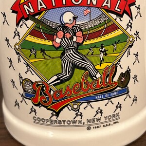 May include: White ceramic mug featuring a baseball theme. The mug has a colorful graphic of a baseball player in a stadium, with the words "NATIONAL BASEBALL HALL OF FAME COOPERSTOWN, NEW YORK" and the year 1987.