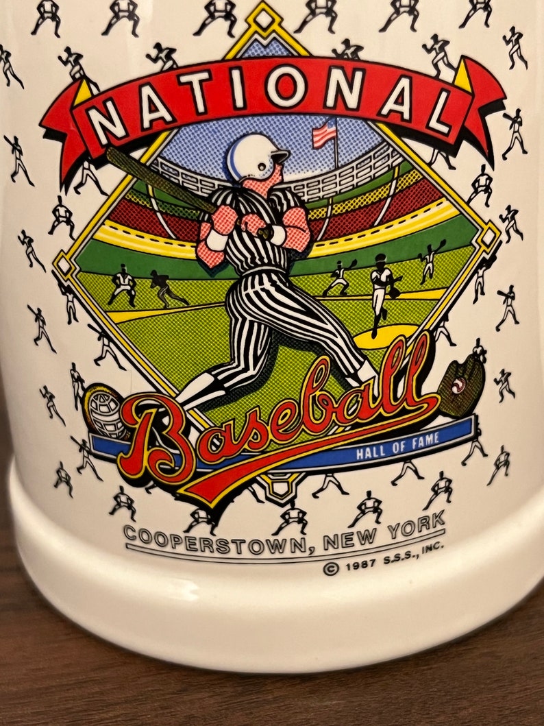 May include: White ceramic mug featuring a baseball theme. The mug has a colorful graphic of a baseball player in a stadium, with the words "NATIONAL BASEBALL HALL OF FAME COOPERSTOWN, NEW YORK" and the year 1987.