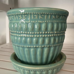 May include: A turquoise ceramic plant pot with a matching saucer. The pot features a glossy finish, a wide rim, and a textured exterior with horizontal bands of raised dots and vertical fluted grooves around the lower section of the vessel.
