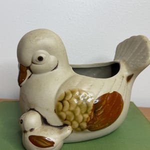 May include: A ceramic planter shaped like a mother bird with a smaller bird attached to the front. The piece is off-white with brown and green painted details on the wings and beak, featuring a hollow opening on the back for plants.