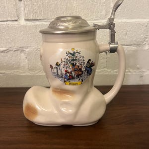 May include: A white ceramic boot-shaped beer stein with a pewter lid and a hinged handle. The stein features a colorful illustration of a Christmas tree and people celebrating. The text "Winter" is printed on the stein.
