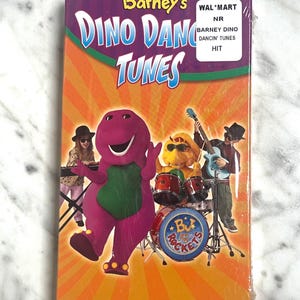 May include: A VHS tape of Barney's Dino Dance Tunes. The cover features Barney the dinosaur with a band, including a drummer, keyboardist, and guitarist. The background is orange with a burst design.