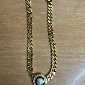 May include: Gold-tone chain necklace with a large circular pendant. The pendant features a central pearl surrounded by a black and navy blue enamel ring, and a border of small clear crystals. The necklace is displayed on a light brown surface.